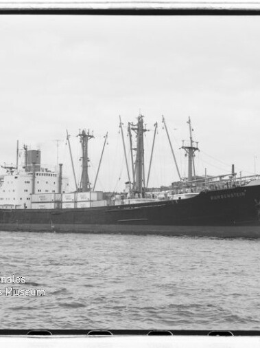 Großes Frachtschiff mit mehreren Ladekränen und Aufbauten liegt am Kai vor Hafenbauten und Uferpromenade.