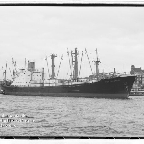 Großes Frachtschiff mit mehreren Ladekränen und Aufbauten liegt am Kai vor Hafenbauten und Uferpromenade.