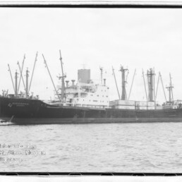 Großes Frachtschiff mit mehreren Ladekränen liegt im Hafen, im Hintergrund ein markanter Kirchturm.