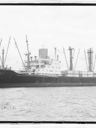 Großes Frachtschiff mit mehreren Ladekränen liegt im Hafen, im Hintergrund ein markanter Kirchturm.