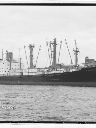 Schwarzweißaufnahme eines großen Frachtschiffs mit mehreren Ladekränen im Hafen vor Uferbebauung; Wasserzeichen des Internationalen Maritimen Museums Hamburg unten links.