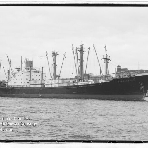 Schwarzweißaufnahme eines großen Frachtschiffs mit mehreren Ladekränen im Hafen vor Uferbebauung; Wasserzeichen des Internationalen Maritimen Museums Hamburg unten links.