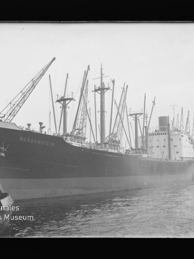 Großes Frachtschiff längsseits am Kai mit mehreren Ladekränen und Masten, schwarz-weiß aufgenommen.