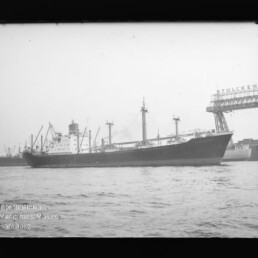 Großes Frachtschiff im Hafen, dahinter Kräne und ein hoher Hafenkran mit teilweise lesbarer Aufschrift.