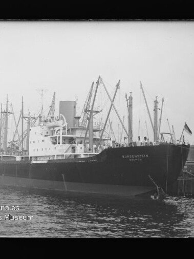 Frachtschiff am Kai mit zahlreichen Hafenkränen, gut sichtbarer Bugaufschrift und Wasser im Vordergrund.