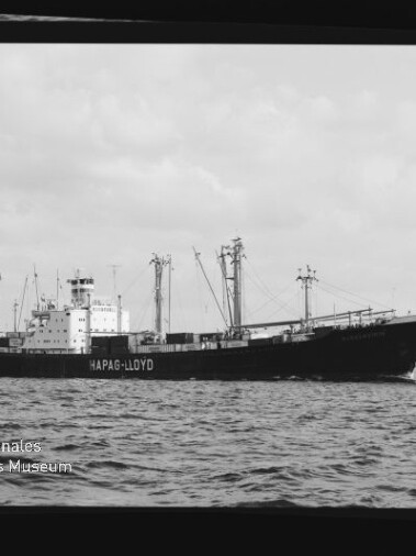 Frachtschiff mit HAPAG-LLOYD-Schriftzug, Ladekräne und Masten auf ruhiger See; Schwarzweißaufnahme mit Museumslogo.