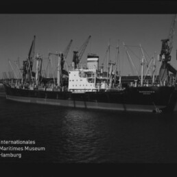Schwarzweißaufnahme eines Frachtschiffs am Kai, umgeben von mehreren Ladekränen im Hafen.