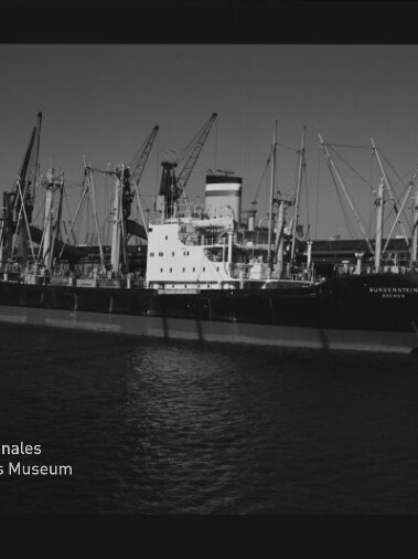 Schwarzweißaufnahme eines Frachtschiffs am Kai, umgeben von mehreren Ladekränen im Hafen.