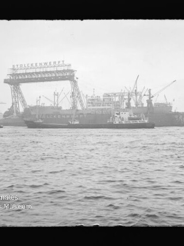 Blick über das Wasser auf eine Werft mit großem Gitterrahmenkran, mehreren Frachtschiffen und zahlreichen Hafenkränen.