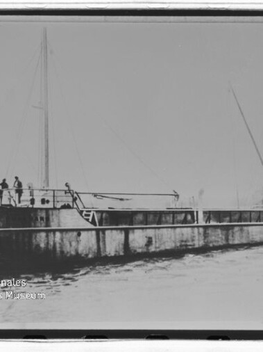 Flaches Frachtschiff mit zwei Masten und mehreren Personen auf dem Deck, fahrend auf dem Wasser.