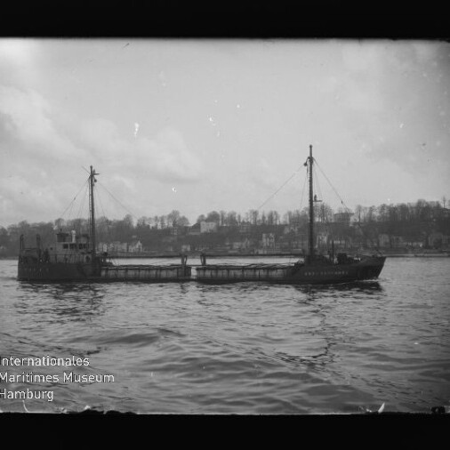 Frachtschiff mit zwei Masten auf ruhigem Wasser vor Ufer mit Häusern; Wasserzeichen des Int. Maritimen Museums Hamburg.