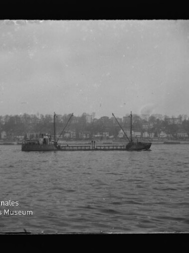 Frachtschiff mit zwei Ladekränen auf ruhigem Wasser, Häuser und Bäume am Ufer im Hintergrund; Museumslogo unten links.