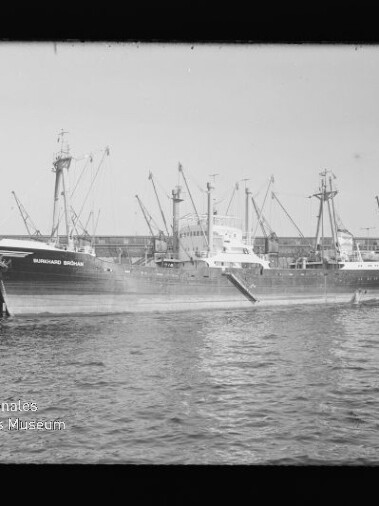 Frachtschiff mit mehreren Ladebäumen liegt am Kai vor Hafenkränen; Wasserzeichen Internationales Maritimes Museum Hamburg links unten.