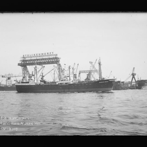 Frachtschiff vor einer Werft mit großen Kränen und markanter Hallenaufschrift im Hintergrund, ruhige See.