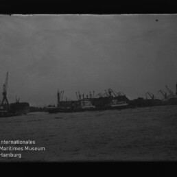 Hafen mit Kränen und Schiffen, Wasser im Vordergrund; Logo des Internationalen Maritimen Museums Hamburg links unten.