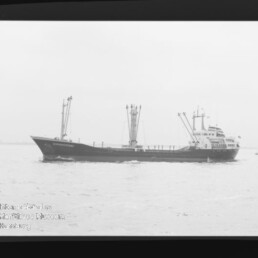 Schwarzweißfoto eines Frachtschiffs mit mehreren Ladekränen auf ruhiger See, kleines Boot am rechten Bildrand.