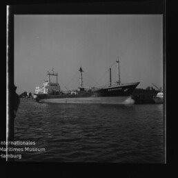 Schwarzweißfoto eines Frachtschiffs im Hafen, Seitenansicht, ruhiges Wasser, Hafenkräne im Hintergrund und Museumslogo unten links.