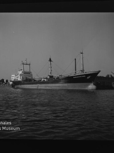 Schwarzweißfoto eines Frachtschiffs im Hafen, Seitenansicht, ruhiges Wasser, Hafenkräne im Hintergrund und Museumslogo unten links.