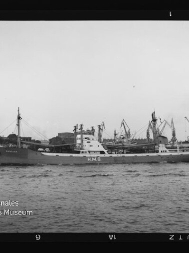 Frachtschiff mit HMG am Rumpf fährt durch einen Industriehafen mit Kränen, daneben ein kleines Schlepperboot.