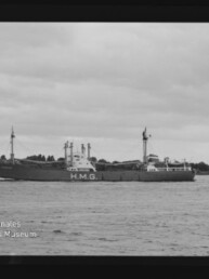 Frachtschiff mit großem H.M.G.-Schriftzug auf dem Rumpf fährt auf ruhiger See vor flachem Ufer, bewölkter Himmel.