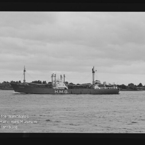 Frachtschiff mit großem H.M.G.-Schriftzug auf dem Rumpf fährt auf ruhiger See vor flachem Ufer, bewölkter Himmel.