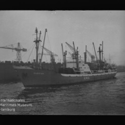 Frachtschiff mit sichtbaren Buchstaben HMG an Bord fährt im Hafen vorbei, im Hintergrund mehrere Hafenkräne.