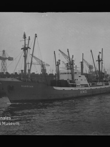 Frachtschiff mit sichtbaren Buchstaben HMG an Bord fährt im Hafen vorbei, im Hintergrund mehrere Hafenkräne.