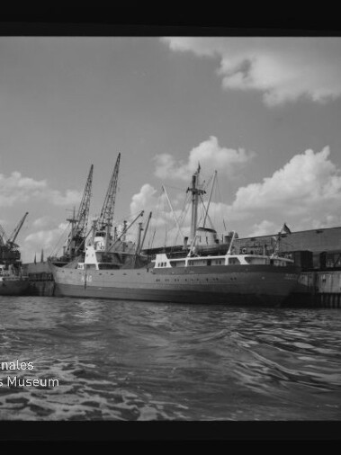 Schiff am Kai mit Hafenkränen, welliges Wasser im Vordergrund und Museumslogo 