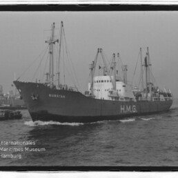 Frachtschiff BURSTAH mit großer Aufschrift H.M.G., begleitet von Schleppern im Hafen vor Hafenkränen.