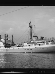 Schwarzweißfoto eines Frachtschiffs mit Aufschrift H.M.G. auf ruhigem Wasser, Ufer im Hintergrund und Museumslogo unten links.