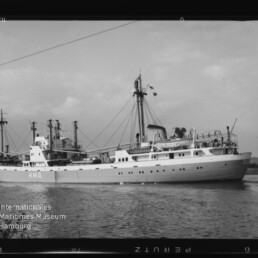 Schwarzweißfoto eines Frachtschiffs mit Aufschrift H.M.G. auf ruhigem Wasser, Ufer im Hintergrund und Museumslogo unten links.