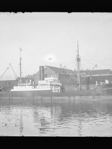 Schwarzweiß-Foto eines Frachtschiffs mit Masten und Aufbauten im Hafen vor Industriegebäuden, Spiegelung im Wasser.