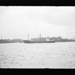 Schwarzweißbild eines Frachtschiffs mit zwei Masten auf dem Wasser vor einem Industriehafen.
