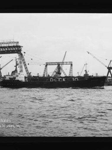 Frachtschiff vor großen Trockendock-Gestellen und Kränen im Hafen, Wasser im Vordergrund, Schriftzug Dock 10 sichtbar.