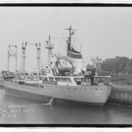 Schwarzweißaufnahme eines Frachtschiffs mit Ladekränen am Kai; unten links sichtbar Internationales Maritimes Museum Hamburg