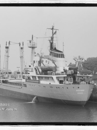 Schwarzweißaufnahme eines Frachtschiffs mit Ladekränen am Kai; unten links sichtbar Internationales Maritimes Museum Hamburg