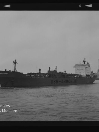 Tanker mit Aufschrift DSR-AMMONIA in Seitenansicht auf dem Wasser vor einem Industriehafen mit zwei hohen Schornsteinen.