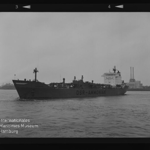 Tanker mit Aufschrift DSR-AMMONIA in Seitenansicht auf dem Wasser vor einem Industriehafen mit zwei hohen Schornsteinen.