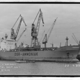 Frachtschiff DSR-AMMONIAK am Kai, umgeben von hohen Hafenkränen und einem kleinen Boot im Vordergrund.