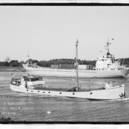 Zwei vorbeifahrende Schiffe auf einem Wasserweg: vorn ein schmales weißes Küstenmotorschiff mit Mast, dahinter ein größeres Frachtschiff; Museumsstempel links unten.