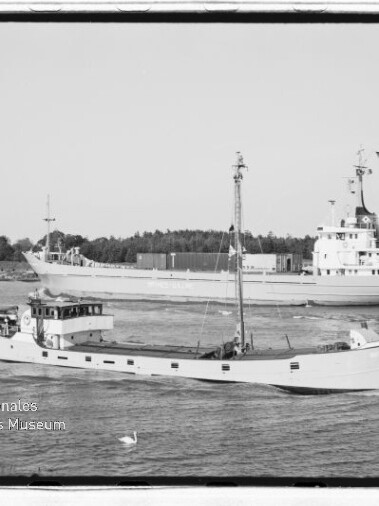 Zwei vorbeifahrende Schiffe auf einem Wasserweg: vorn ein schmales weißes Küstenmotorschiff mit Mast, dahinter ein größeres Frachtschiff; Museumsstempel links unten.