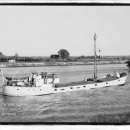 Kleines Frachtschiff auf Kanal in Schwarzweiß, Ufer im Hintergrund und Museumssignet Internationales Maritimes Museum Hamburg.