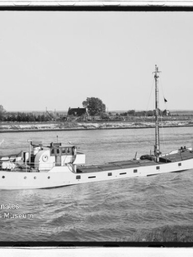 Kleines Frachtschiff auf Kanal in Schwarzweiß, Ufer im Hintergrund und Museumssignet Internationales Maritimes Museum Hamburg.