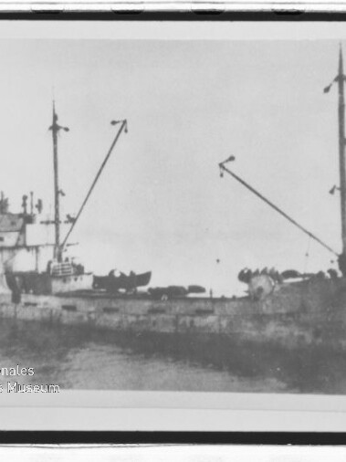 Altes Frachtschiff mit zwei Masten und schrägen Ladebäumen, sichtbares Museumswasserzeichen unten links.