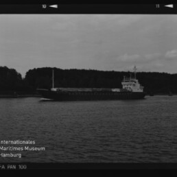 Frachtschiff auf ruhigem Wasser vor bewaldetem Ufer; Schwarzweiß-Filmrahmen mit Stempel 