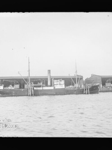 Dampfschiff mit Schornstein und Masten am Kai festgemacht, vor langen Lagerhallen, ruhige Wasserfläche im Vordergrund.