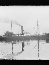 Schwarzweißaufnahme eines dampfgetriebenen Frachtschiffs mit rauchendem Schornstein und Spiegelung auf ruhigem Wasser.