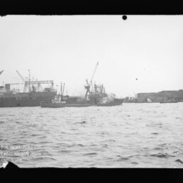 Blick übers Wasser auf Hafen mit Frachtschiffen, Kränen und Schleppern; Logo 'Internationales Maritimes Museum Hamburg'.