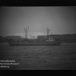 Frachtschiff mit Masten und Aufbauten vor einer Hafen-Silhouette, ruhige See; Museumslogo unten links sichtbar.