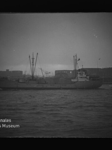 Frachtschiff mit Masten und Aufbauten vor einer Hafen-Silhouette, ruhige See; Museumslogo unten links sichtbar.